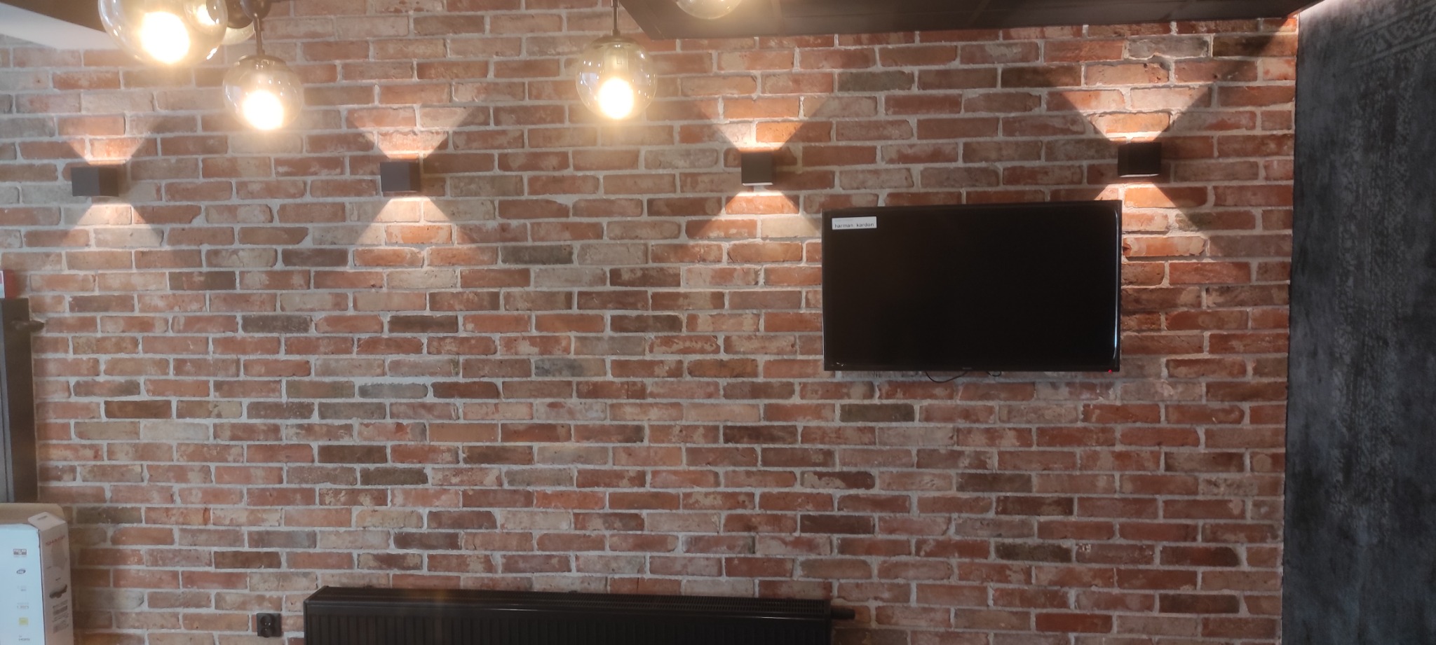 Ściana z cegły klinkierowej z zamontowanym telewizorem i czarnymi kinkietami emitującymi ciepłe światło, widoczne również wiszące lampy z żarówkami Edison.