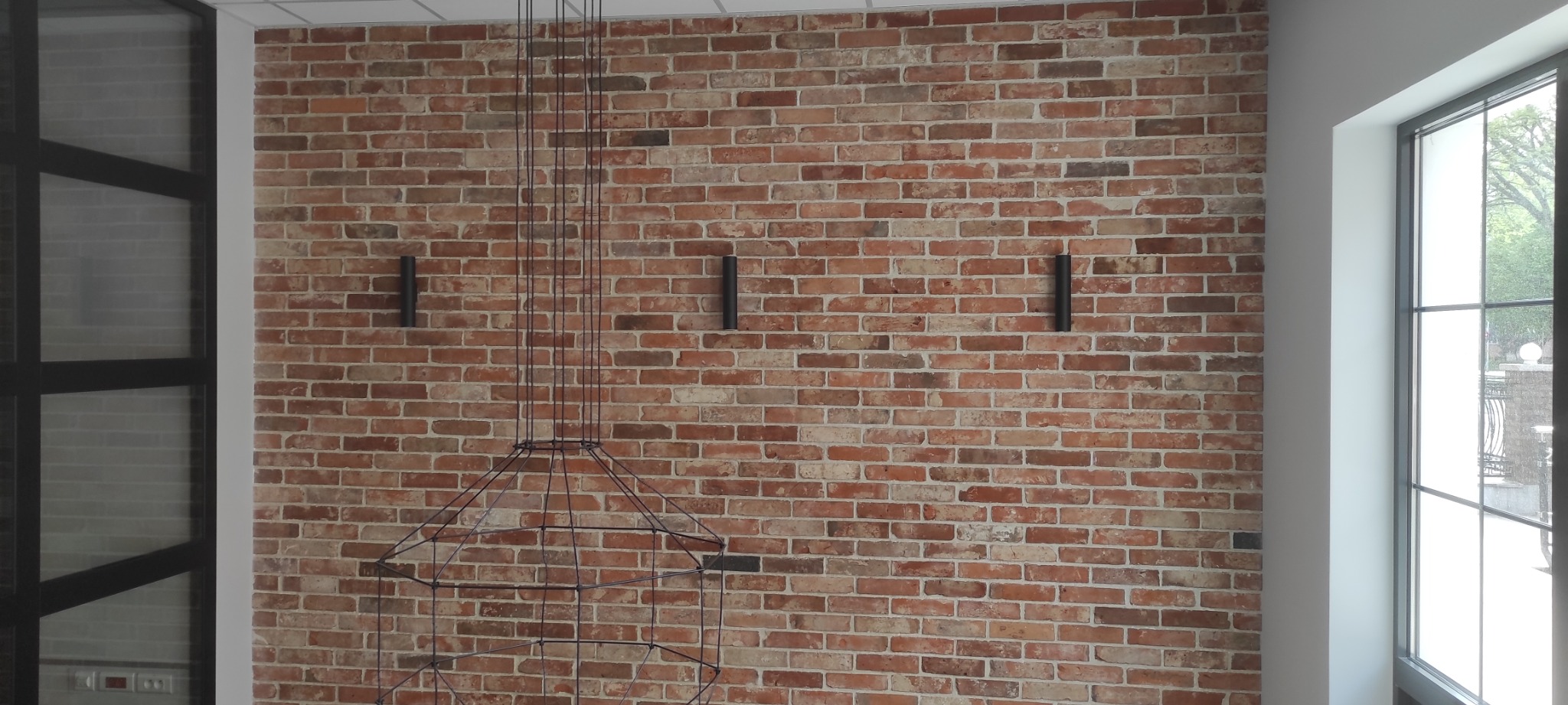 Ściana z czerwonej cegły z trzema czarnymi kinkietami i metalową konstrukcją lampy wiszącej bez klosza.