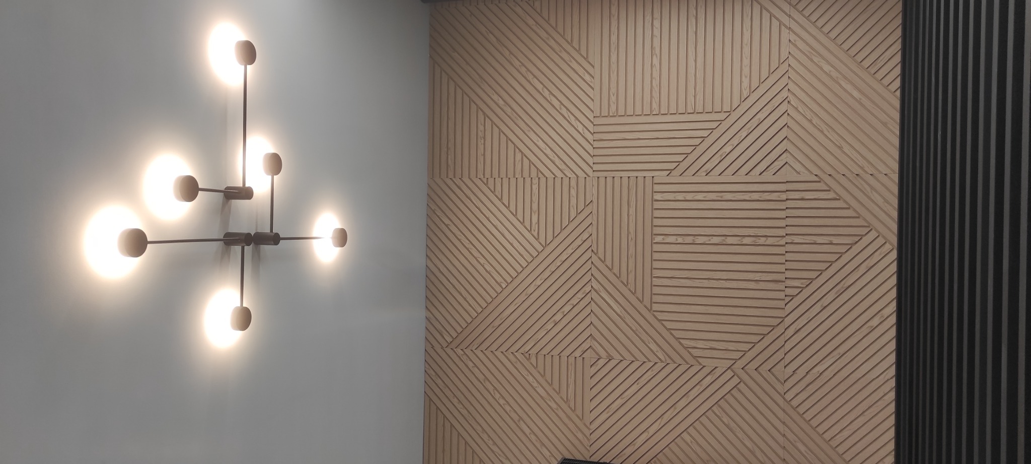 Nowoczesna lampa ścienna z sześcioma okrągłymi źródłami światła na tle ściany z drewnianymi panelami o geometrycznym wzorze.