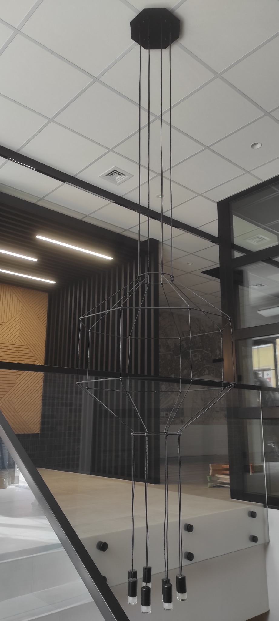 Nowoczesna lampa wisząca z geometrycznym kloszem, zamontowana w biurze z drewnianą ścianą i przeszklonymi drzwiami.