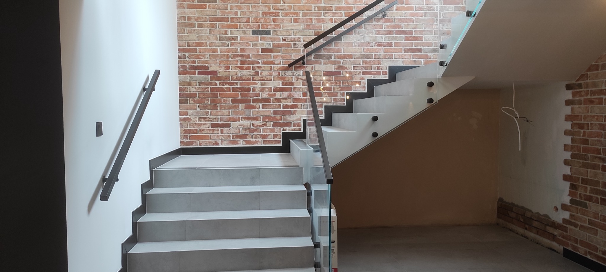 Widok na nowoczesne schody z szarymi stopniami, szklanymi balustradami i czarnymi poręczami w Elblągu, z fragmentem ściany z cegły klinkierowej w tle oraz widocznym wystającym przewodem elektrycznym.