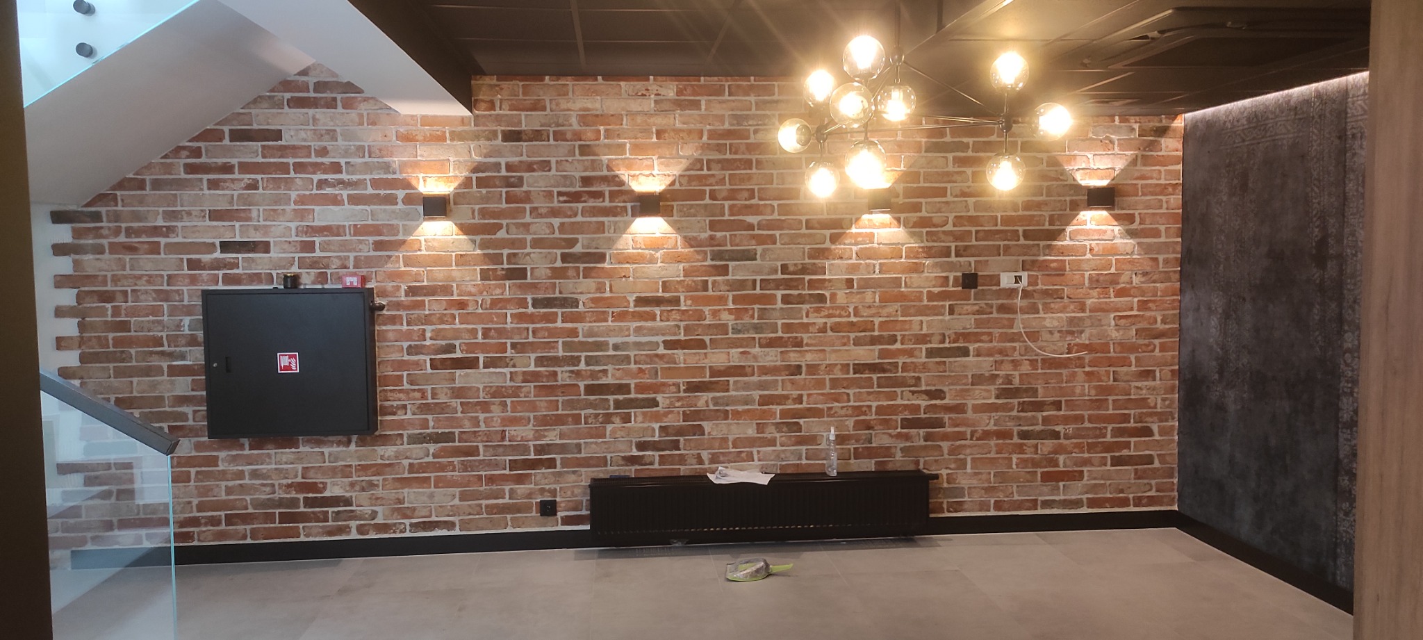 Wnętrze z ceglaną ścianą, ozdobnymi kinkietami i żyrandolem, widoczna szafka elektryczna i wystający przewód z gniazdka.