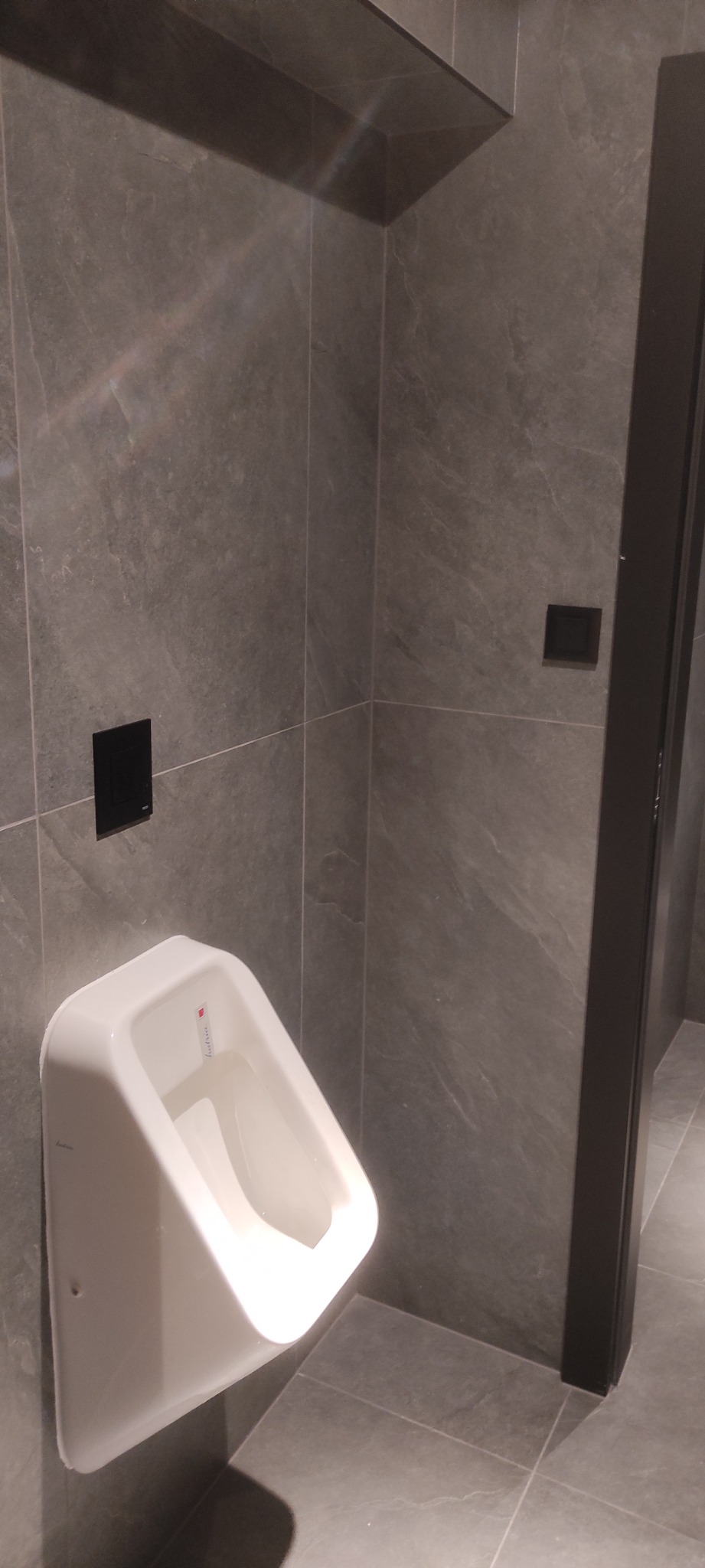 Biały pisuar zamontowany na szarej, marmurkowej ścianie, z czarnym przyciskiem spłukującym i czarną ramą drzwi po prawej stronie.