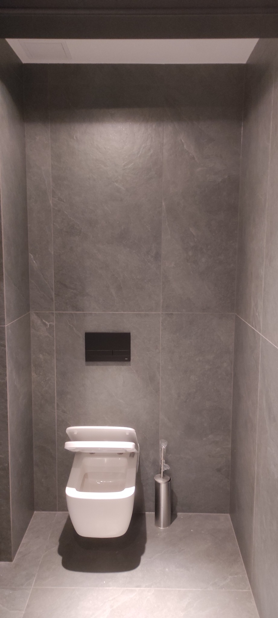 Nowoczesna toaleta podwieszana z czarnym przyciskiem spłukującym na szarej, kamiennej ścianie, uzupełniona o szczotkę do WC w srebrnym pojemniku.