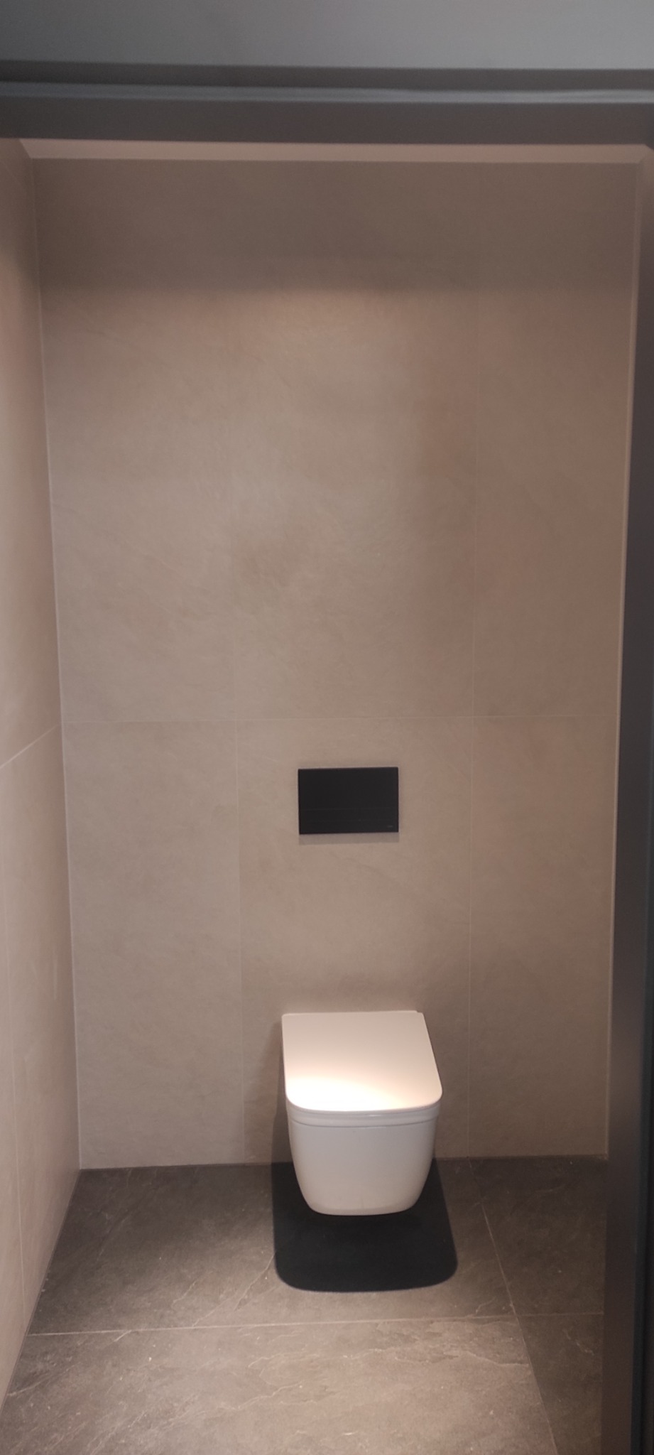 Nowoczesna toaleta podwieszana z czarnym przyciskiem spłukującym, zamontowana na beżowej ścianie wyłożonej płytkami, z szarą podłogą z płytek imitujących kamień.