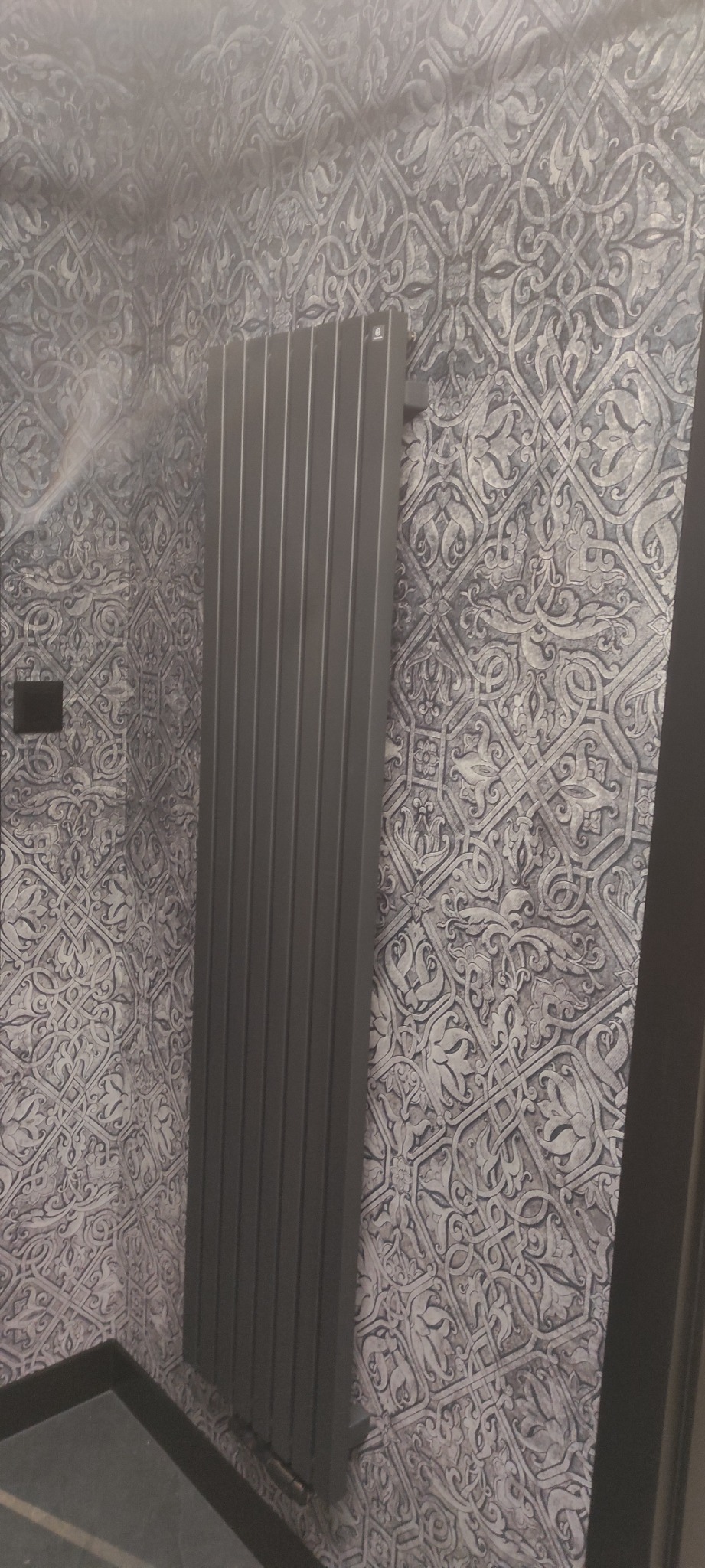 Pionowy grzejnik panelowy w kolorze grafitowym zamontowany na ścianie pokrytej tapetą w marokańskie wzory, widok z niskiej perspektywy.