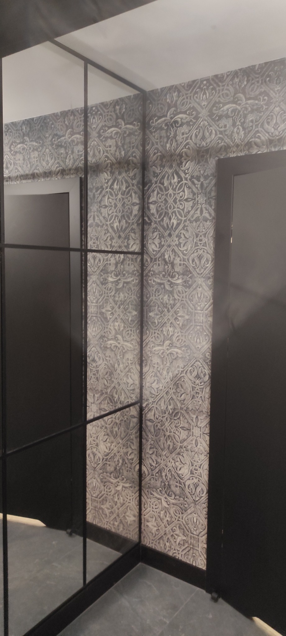 Czarno-biała ściana z wzorzystą tapetą i czarnymi drzwiami, widziana przez szklaną ściankę działową z czarnymi profilami.