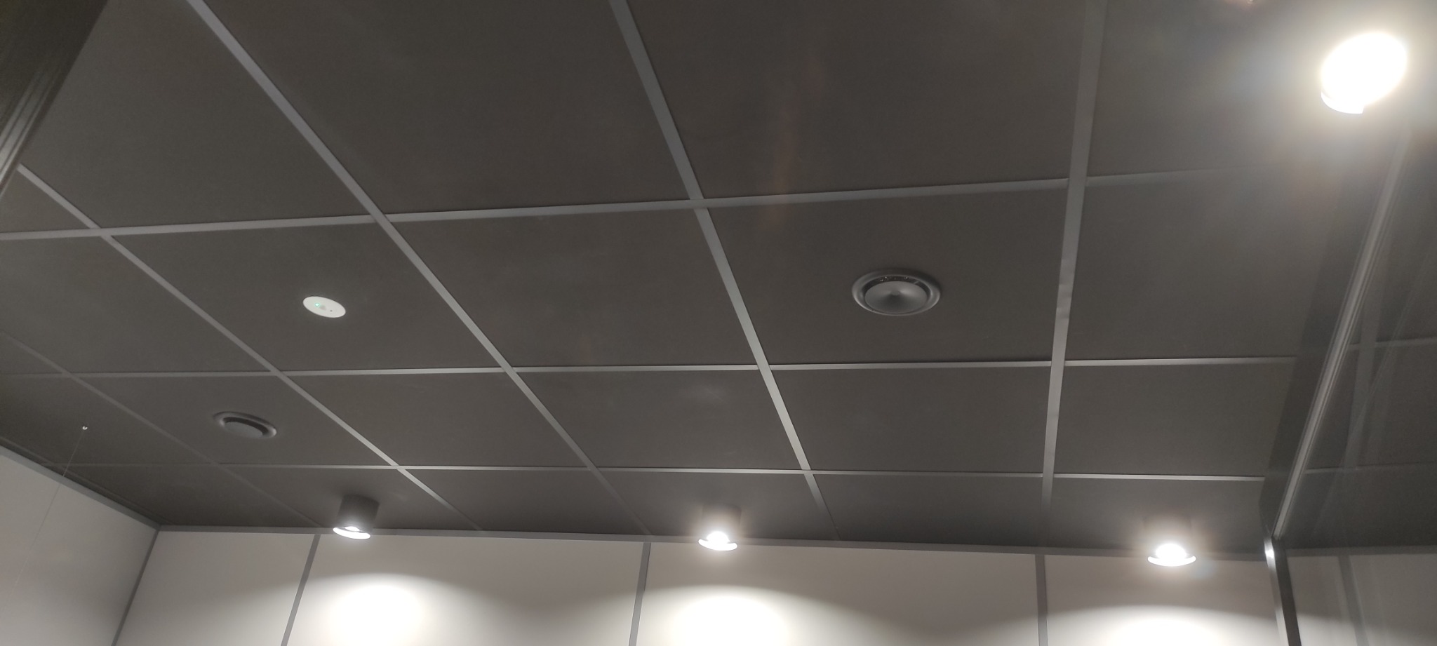 Sufit podwieszany z widocznymi punktami świetlnymi i kratką wentylacyjną, oświetlony z kilku stron, z widocznymi elementami konstrukcyjnymi