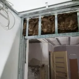 Ocieplenie poddasza wełną mineralną w metalowej konstrukcji szkieletowej, widoczne kable i fragmenty ściany w trakcie remontu.