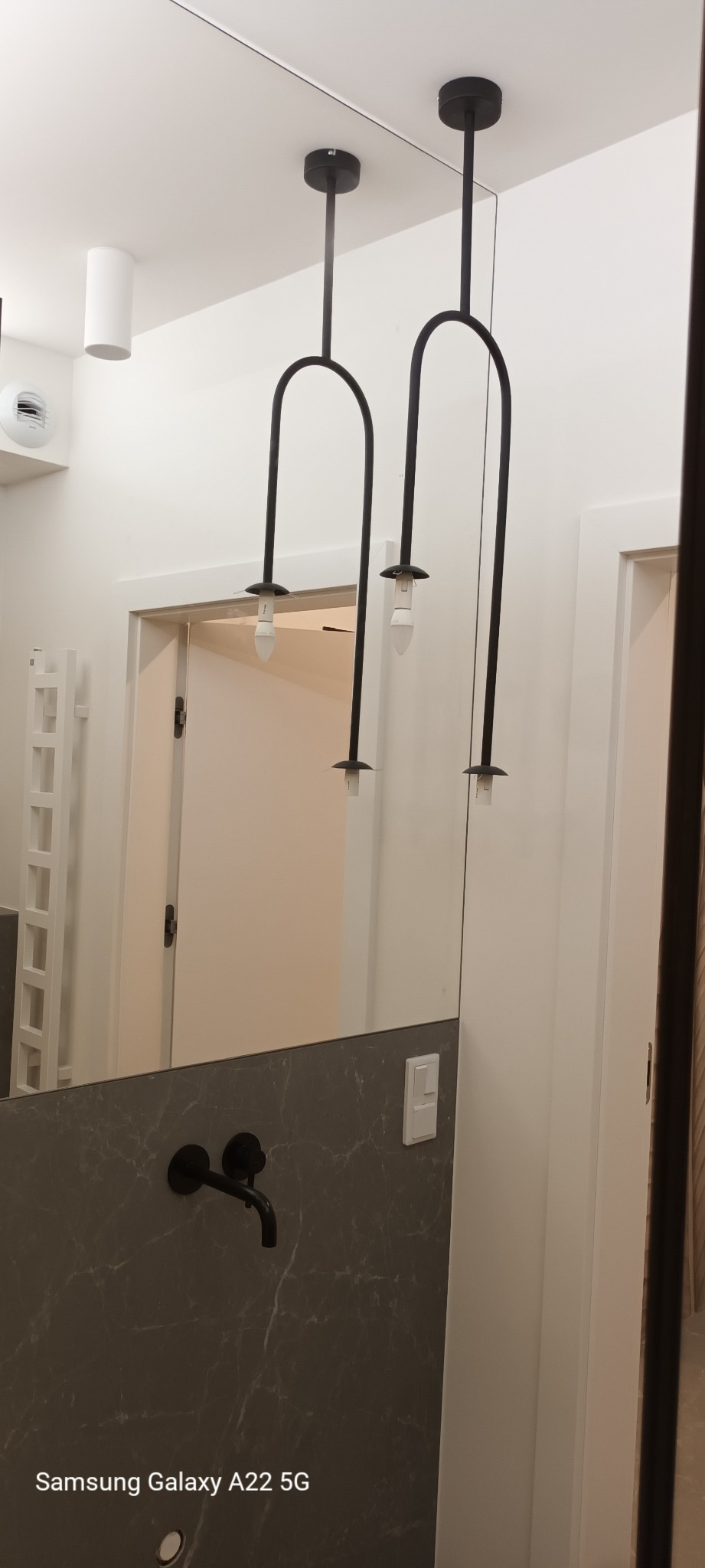 Nowoczesna łazienka z szarą marmurową umywalką, czarną armaturą i designerskim oświetleniem sufitowym w kształcie łuków, widoczna w lustrze.