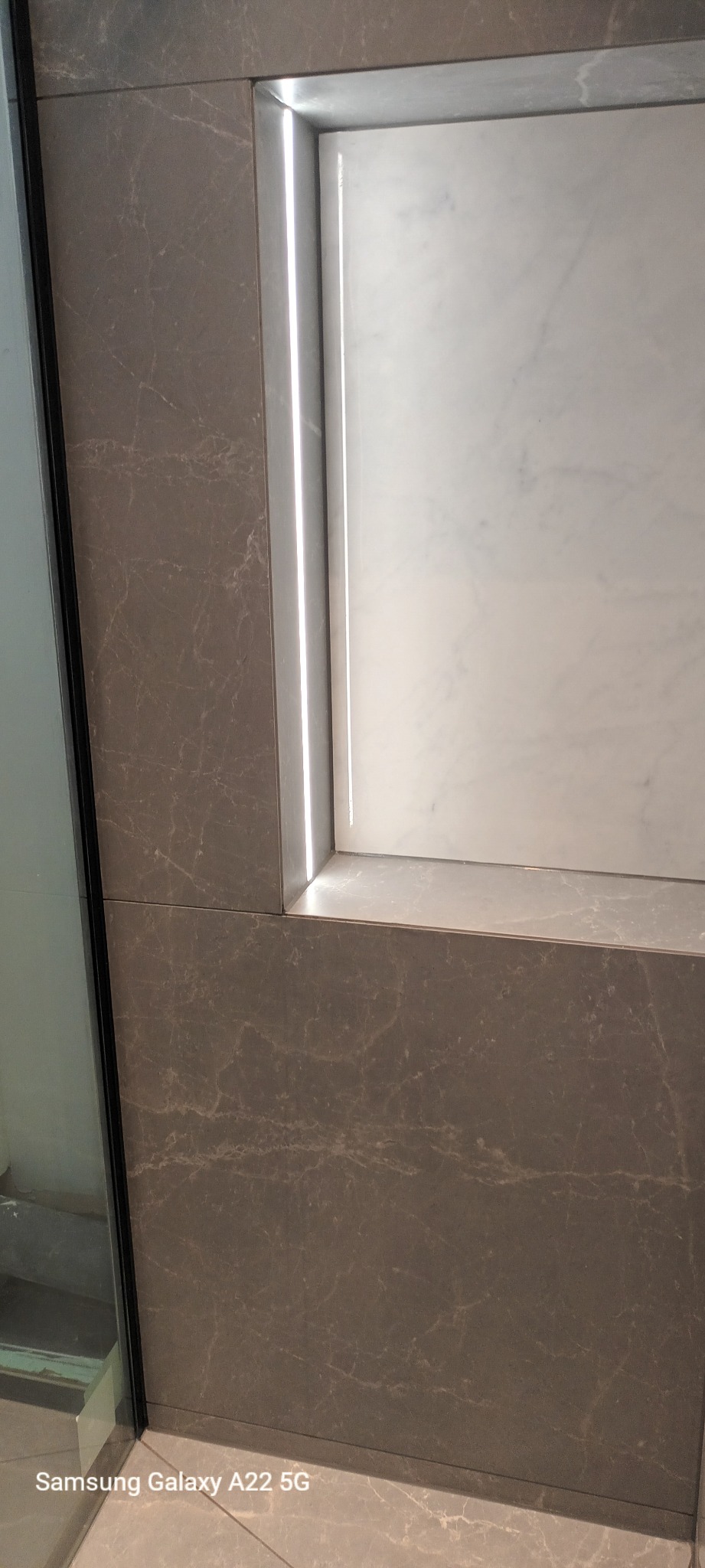 Szara marmurowa ściana z wnęką wyłożoną białym marmurem, oświetloną listwą LED, widok z dołu.