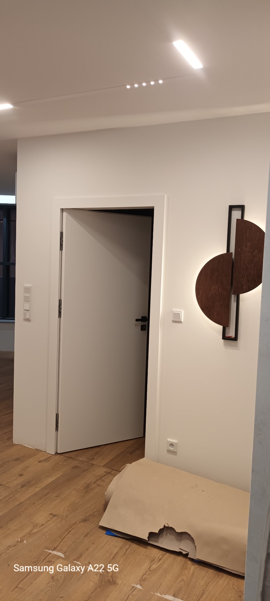 Wnętrze mieszkania w trakcie prac wykończeniowych: widoczna biała ościeżnica z otwartymi białymi drzwiami, drewniana podłoga zabezpieczona kartonem, nowoczesna lampa ścienna z drewnianymi...