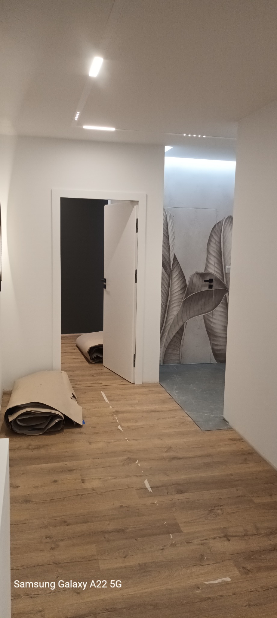 Korytarz w trakcie prac wykończeniowych: położona drewnopodobna podłoga, widoczne rolki z zabezpieczeniem podłogi, drzwi wewnętrzne z czarną ścianą w tle oraz fragment ściany z motywem roślinnym...
