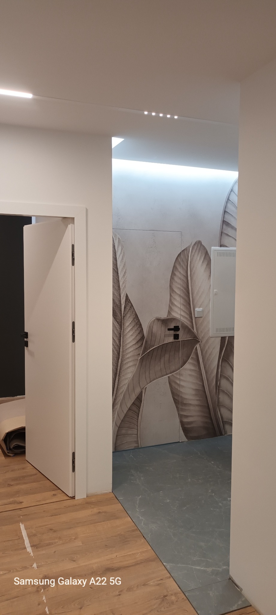 Wnętrze mieszkania po remoncie: otwarta biała drzwi, na ścianie fototapeta z motywem liści bananowca, szara podłoga imitująca marmur, widoczne listwy przypodłogowe.