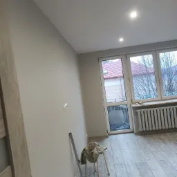 Świeżo pomalowane na szaro ściany w pokoju z drewnopodobną podłogą, widoczny grzejnik, okno i drabina malarska oparta o ścianę. Na krześle leży płachta malarska.