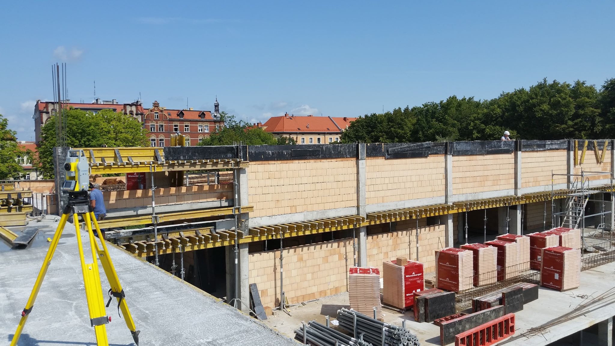 Żółty teodolit na budowie, w tle mur z pustaków ceramicznych Wienerberger, budynek i drzewa.