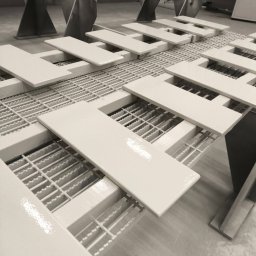 Łukasz Perliceusz - Białe, pomalowane elementy na metalowej kratownicy w hali produkcyjnej. Świeżo nałożona farba tworzy błyszczącą powierzchnię. Ujęcie z perspektywy.