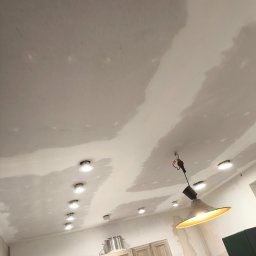 Łukasz Perliceusz - Nisko kątowe ujęcie sufitu w trakcie prac wykończeniowych, widoczne gładzie i zamontowane oświetlenie punktowe. W tle zabudowa kuchenna.