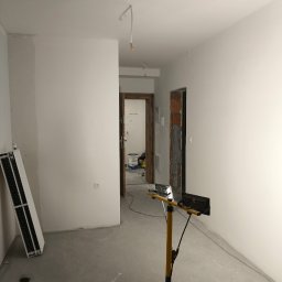 Łukasz Perliceusz - Wnętrze w trakcie remontu: białe ściany po gładzi, widoczna lampa budowlana, grzejnik i narzędzia malarskie. Przygotowanie do malowania w jasnym pomieszczeniu.