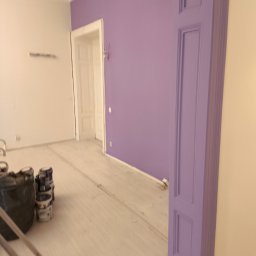 Łukasz Perliceusz - Pomieszczenie po malowaniu ścian na fioletowo, widoczne puszki z farbą i drabina malarska. Wykończone listwy przypodłogowe i ozdobne elementy architektoniczne.
