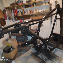 Łukasz Perliceusz - Czarna, metalowa rama motocyklowa po malowaniu, ustawiona w warsztacie. Widoczne półki z narzędziami i akcesoriami w tle. Świeża, błyszcząca powłoka lakiernicza.