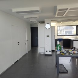 Łukasz Perliceusz - Wnętrze po malowaniu: białe ściany, sufit z panelami, ciemna podłoga. Widoczny stół z narzędziami malarskimi i drabina. Minimalistyczny design, nowoczesne oświetlenie.