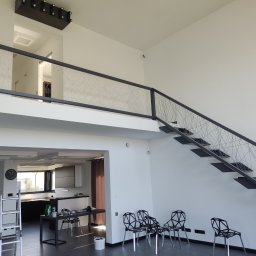 Łukasz Perliceusz - Wnętrze domu po malowaniu: białe ściany, schody z metalową balustradą, widoczna kuchnia. Na podłodze krzesła i akcesoria malarskie.