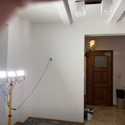 Łukasz Perliceusz - Świeżo pomalowane białe ściany i sufit z ozdobnymi belkami, oświetlone statywem z trzema lampami LED. Widoczne wiadro farby i drabina, w tle drewniane drzwi.