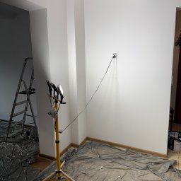Łukasz Perliceusz - Jasne pomieszczenie z gładkimi, białymi ścianami, oświetlone lampą roboczą na statywie. Podłoga zabezpieczona folią malarską, w tle drabina.