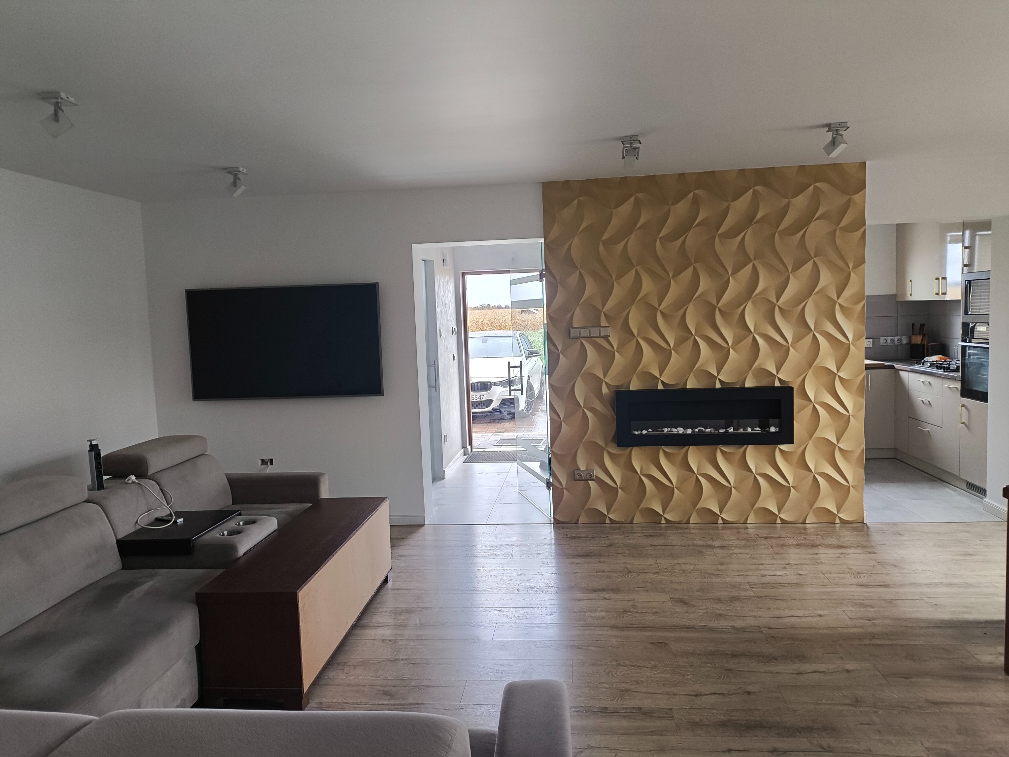 Nowoczesny salon z dekoracyjną ścianą 3D w odcieniach beżu, czarnym kominkiem elektrycznym i drewnianą podłogą. Widoczny fragment sofy i kuchni w tle.