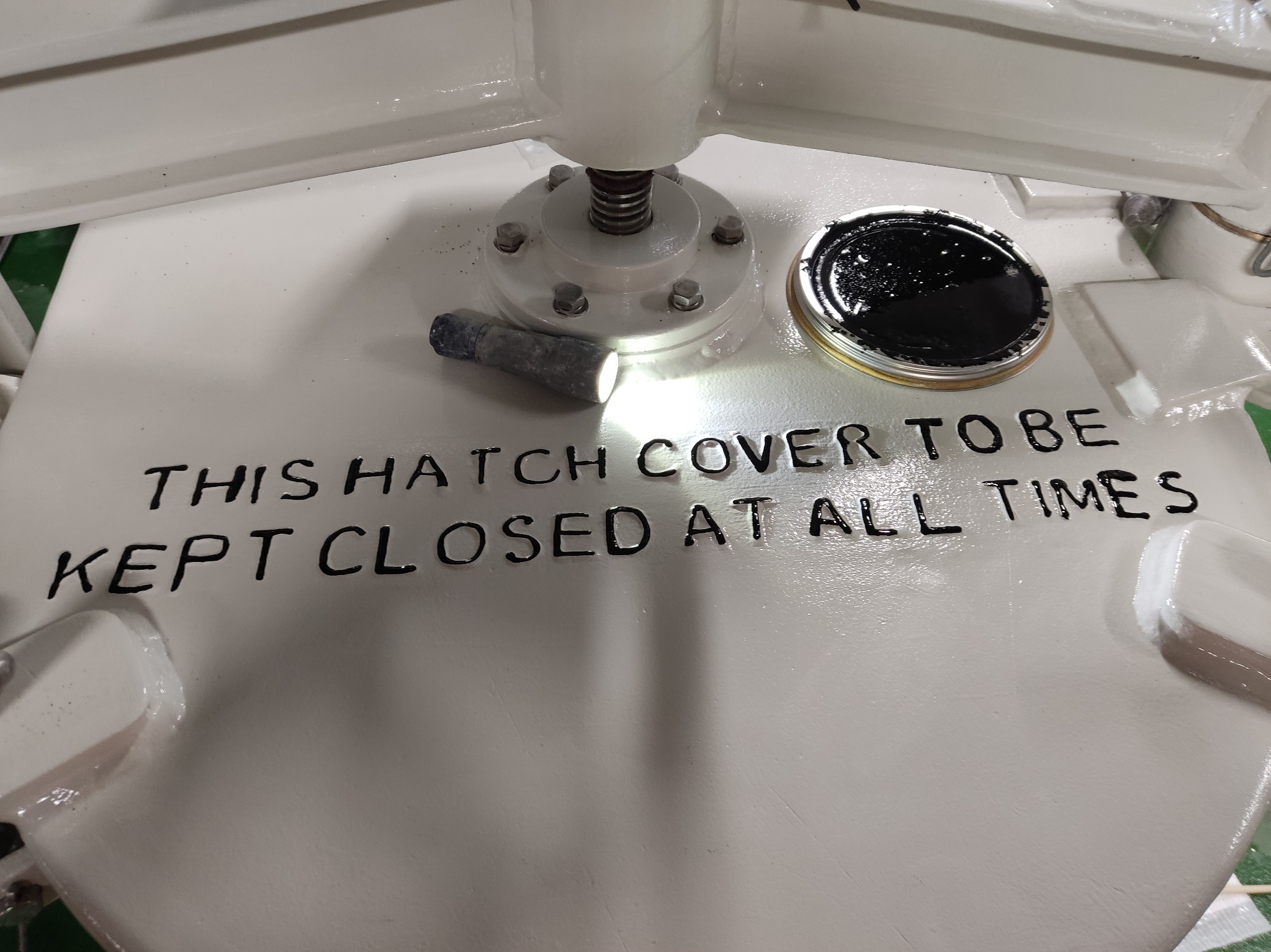 Biała pokrywa włazu z napisem 'THIS HATCH COVER TO BE KEPT CLOSED AT ALL TIMES', obok puszka z czarną farbą i latarka.