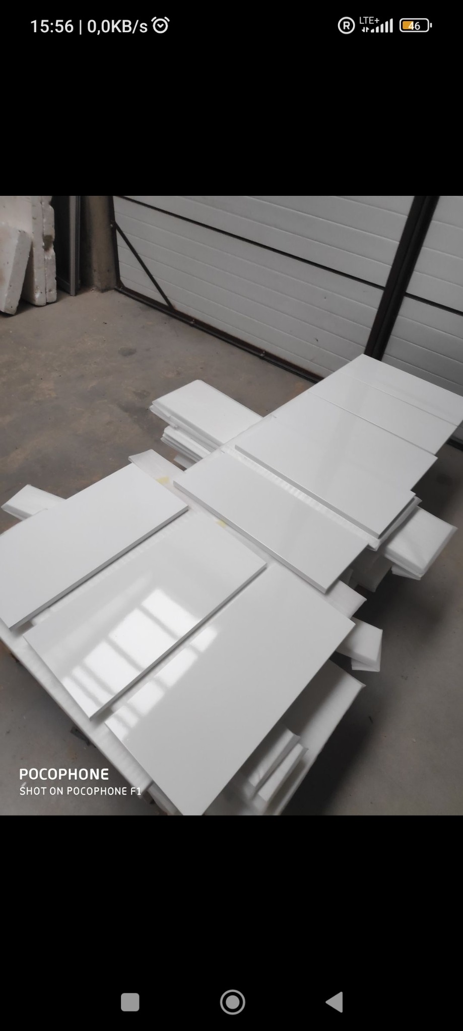 Białe, błyszczące płyty ułożone w stosy na podłodze w magazynie, gotowe do montażu. Widoczna brama garażowa w tle.