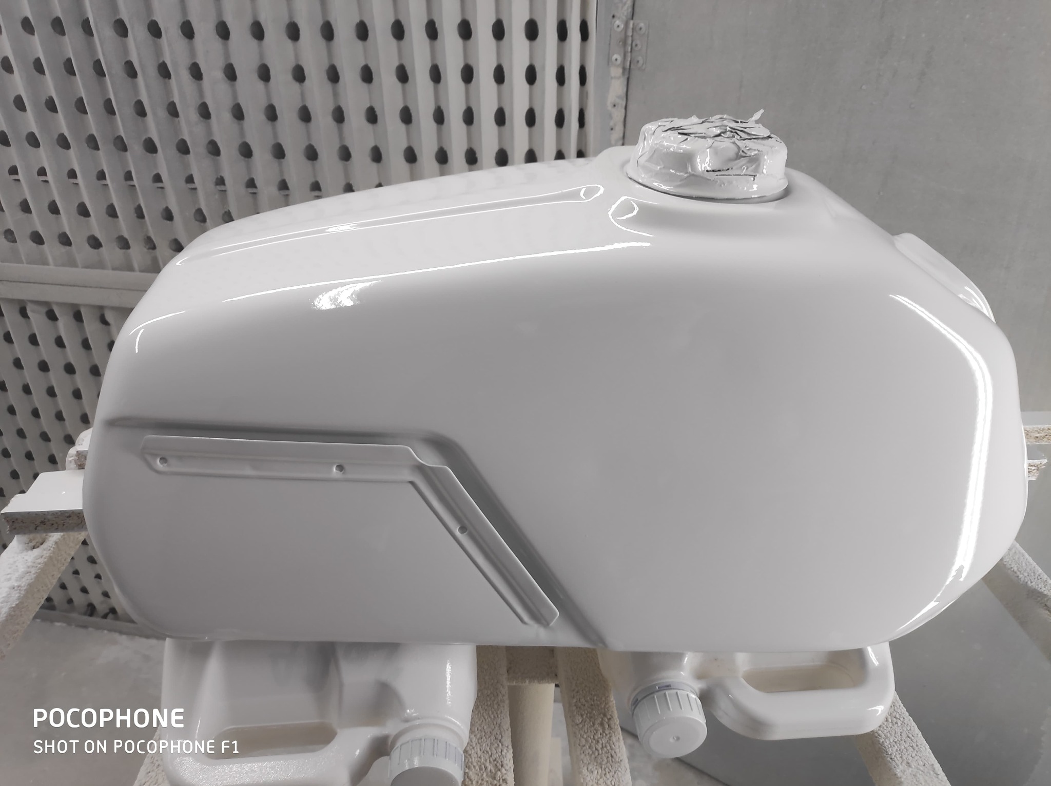 Biały, świeżo pomalowany zbiornik paliwa motocykla w kabinie lakierniczej, z widocznymi detalami i połyskiem lakieru. Ochrona na wlewie paliwa.