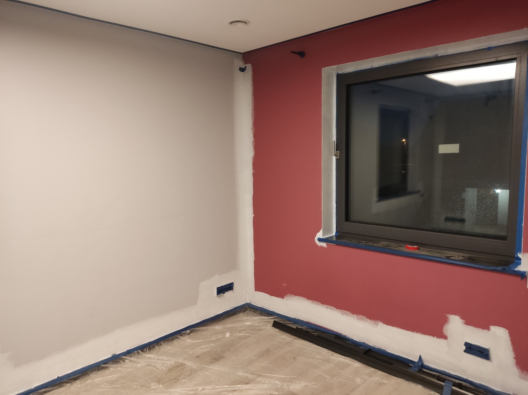 Pokój w trakcie malowania: jedna ściana szara, druga bordowa, zabezpieczone taśmą malarską. Widoczne niedomalowania i folia ochronna na podłodze.