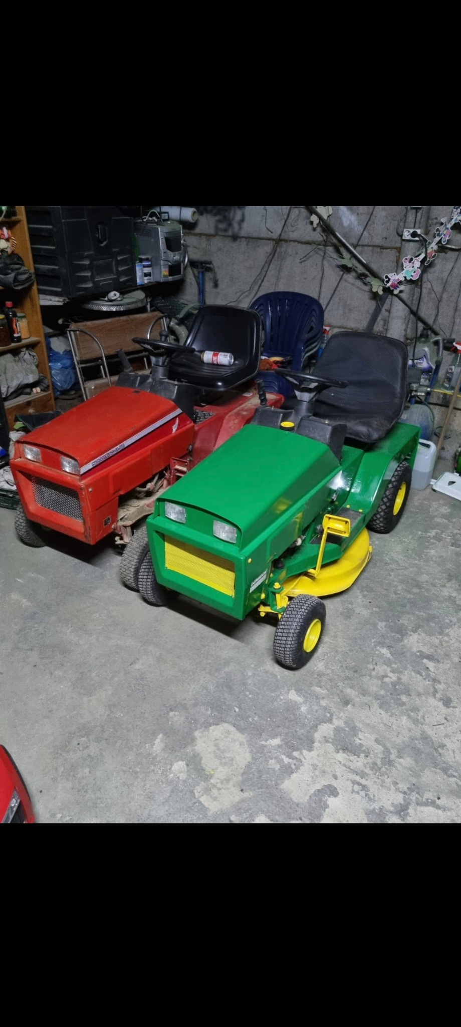 Dwa traktorki do koszenia trawy: czerwony i zielony z żółtymi akcentami, stojące w garażu. Na czerwonym puszka. Widoczne regały i inne przedmioty w tle.