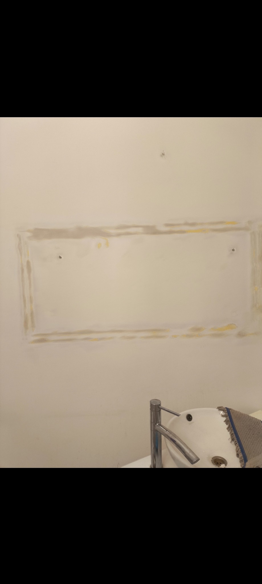 Ściana z wygładzonym prostokątnym obszarem, przygotowana pod montaż lustra lub szafki. Widoczna umywalka z baterią. Ślady po gładzi na ścianie.