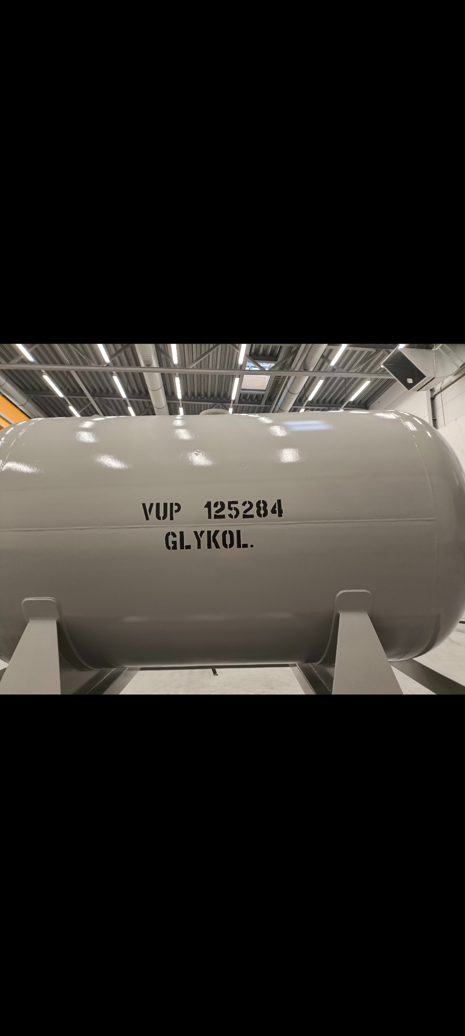 Szary, cylindryczny zbiornik na glikol z czarnym napisem 'VUP 125284 GLYKOL' w hali przemysłowej, oświetlony lampami sufitowymi.