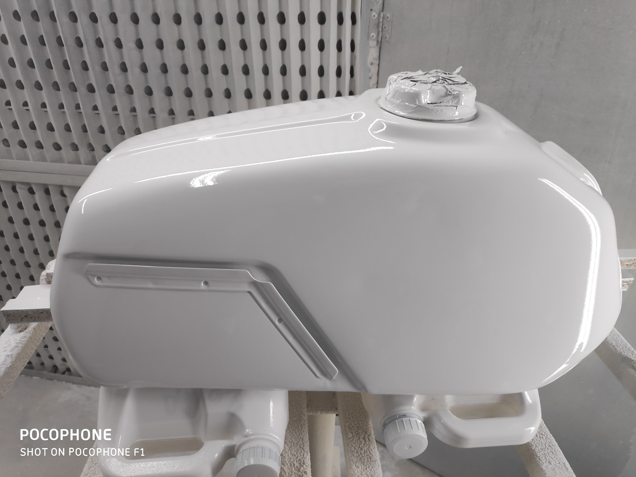 Biały, błyszczący zbiornik paliwa motocykla po lakierowaniu, umieszczony na stojaku w lakierni. Widoczne detale, takie jak wlew paliwa zabezpieczony folią.