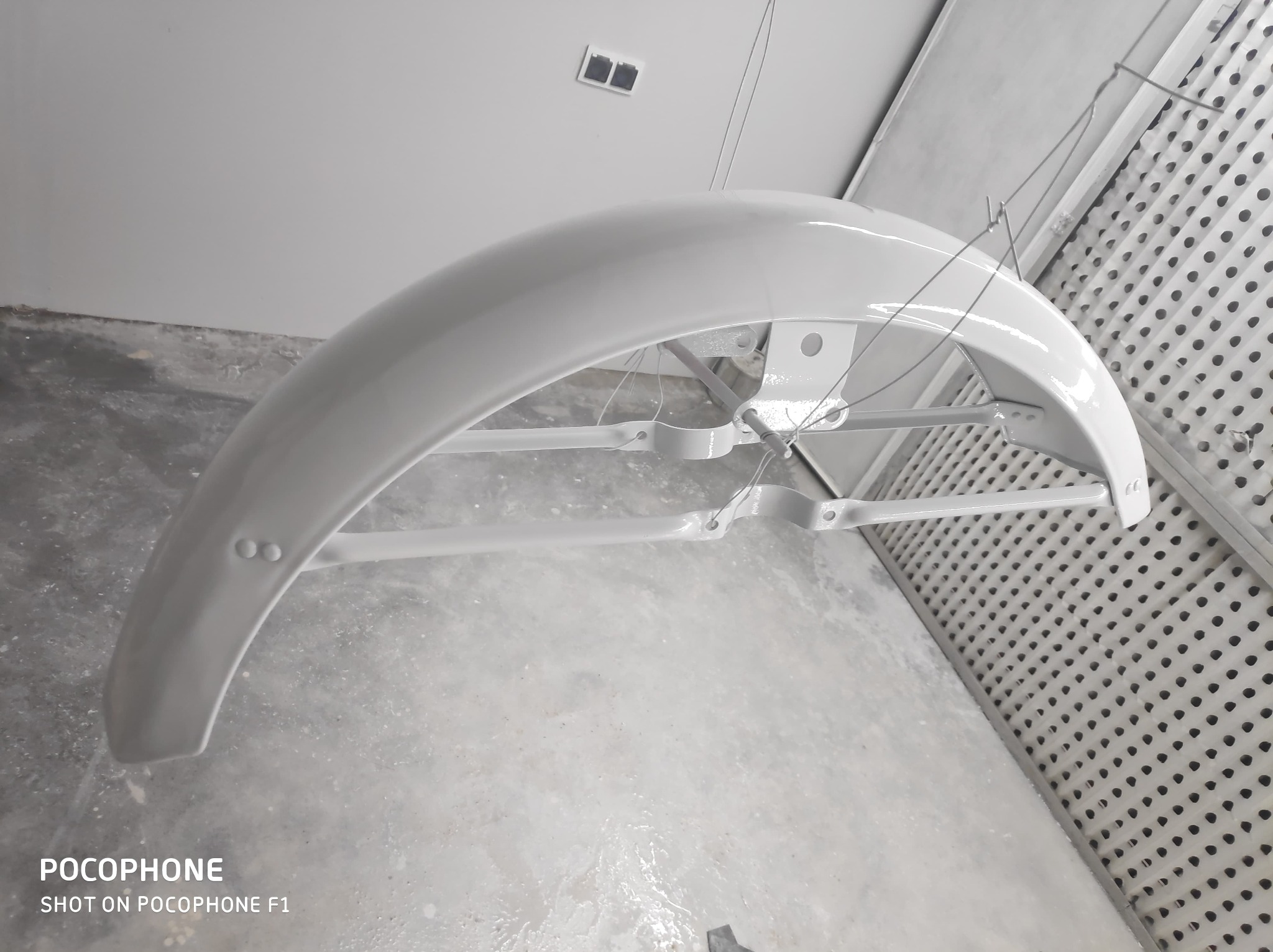 Błotnik rowerowy po lakierowaniu na biało, zawieszony w lakierni. Widoczne detale mocowania i świeża powłoka lakiernicza. Tło stanowią ściany lakierni z kratką wentylacyjną.