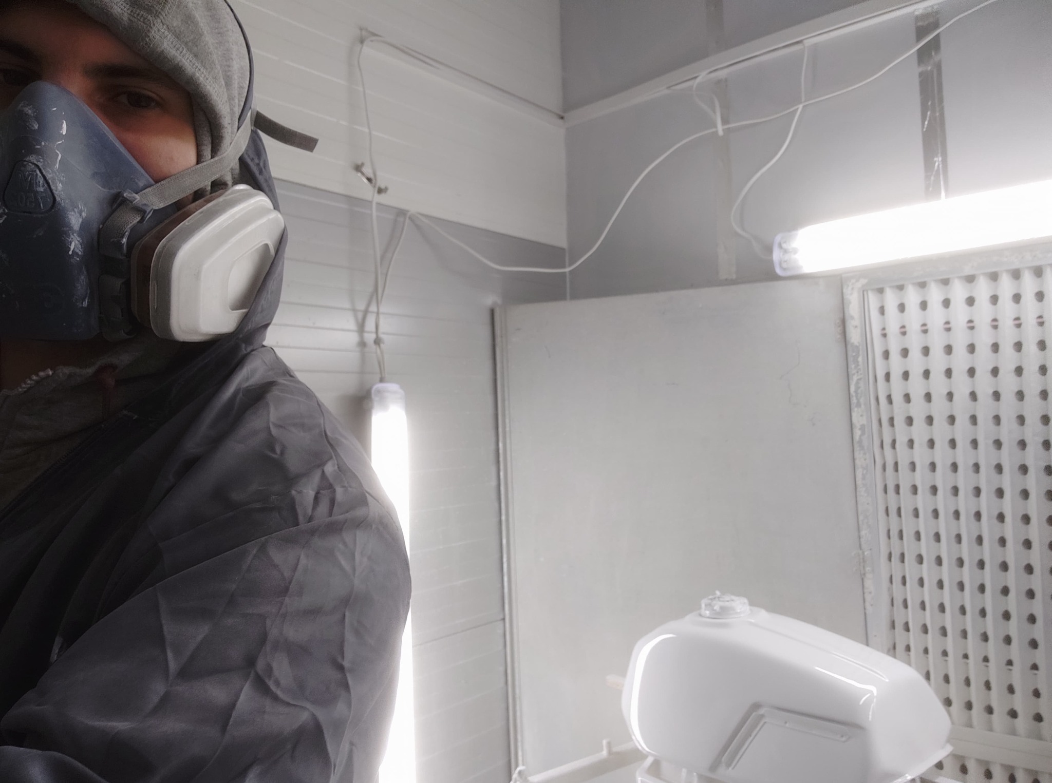 Lakiernik w masce ochronnej, w trakcie lakierowania białego zbiornika paliwa w kabinie lakierniczej z oświetleniem jarzeniowym. Widoczny filtr powietrza.