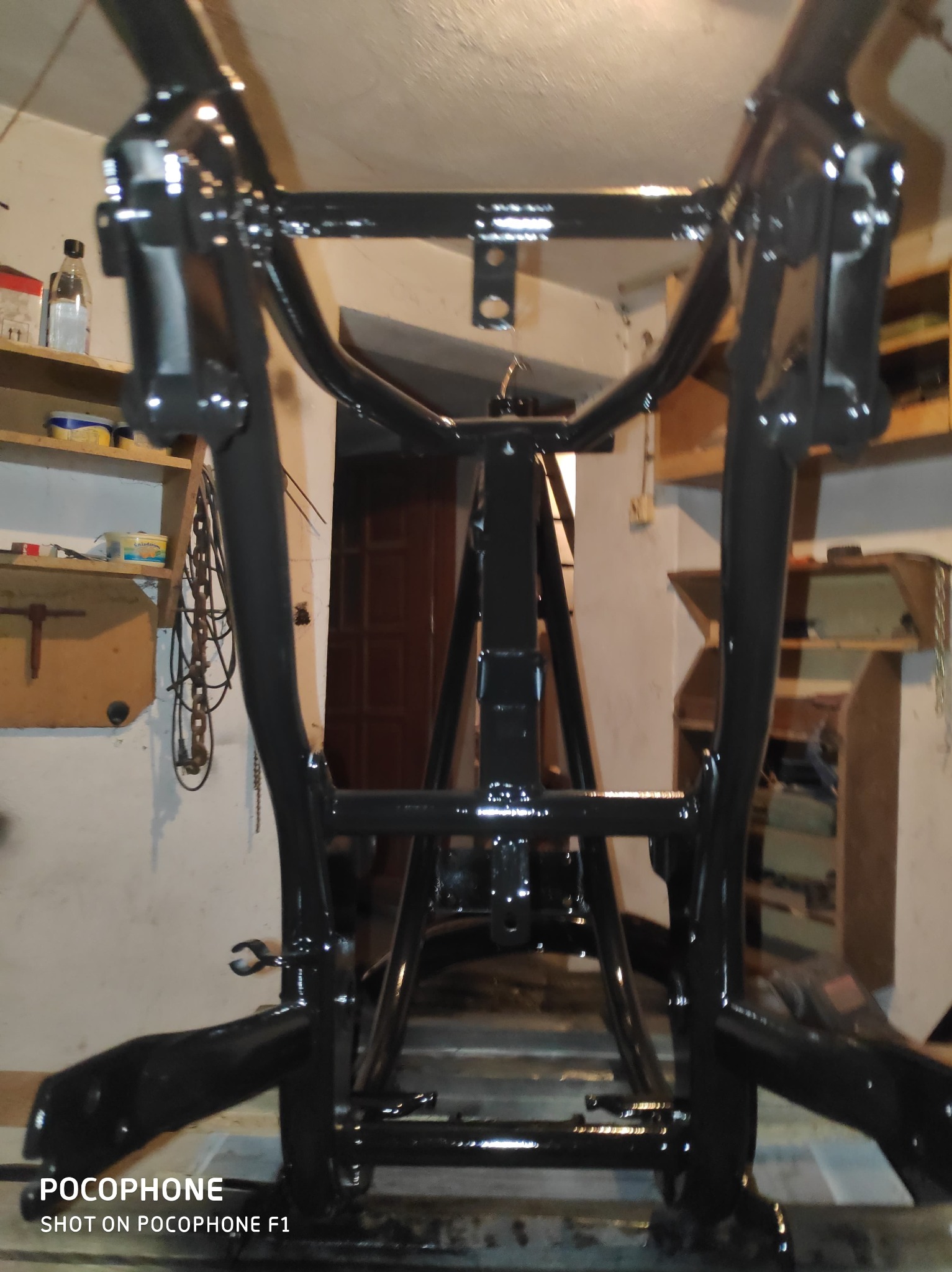 Błyszcząca, czarna rama motocykla po malowaniu, widoczna z bliska w warsztacie. Elementy ramy odbijają światło, ukazując gładką powierzchnię. W tle półki z narzędziami.