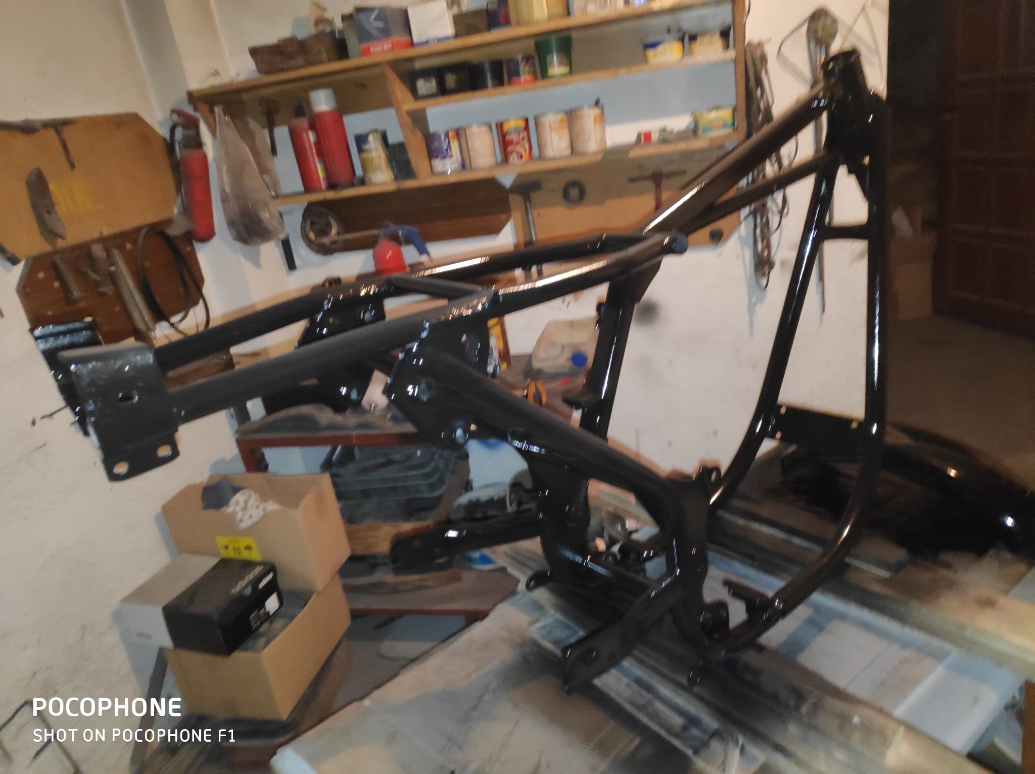 Czarna, metalowa rama motocyklowa po malowaniu, ustawiona w warsztacie. Widoczne półki z narzędziami i akcesoriami w tle. Świeża, błyszcząca powłoka lakiernicza.