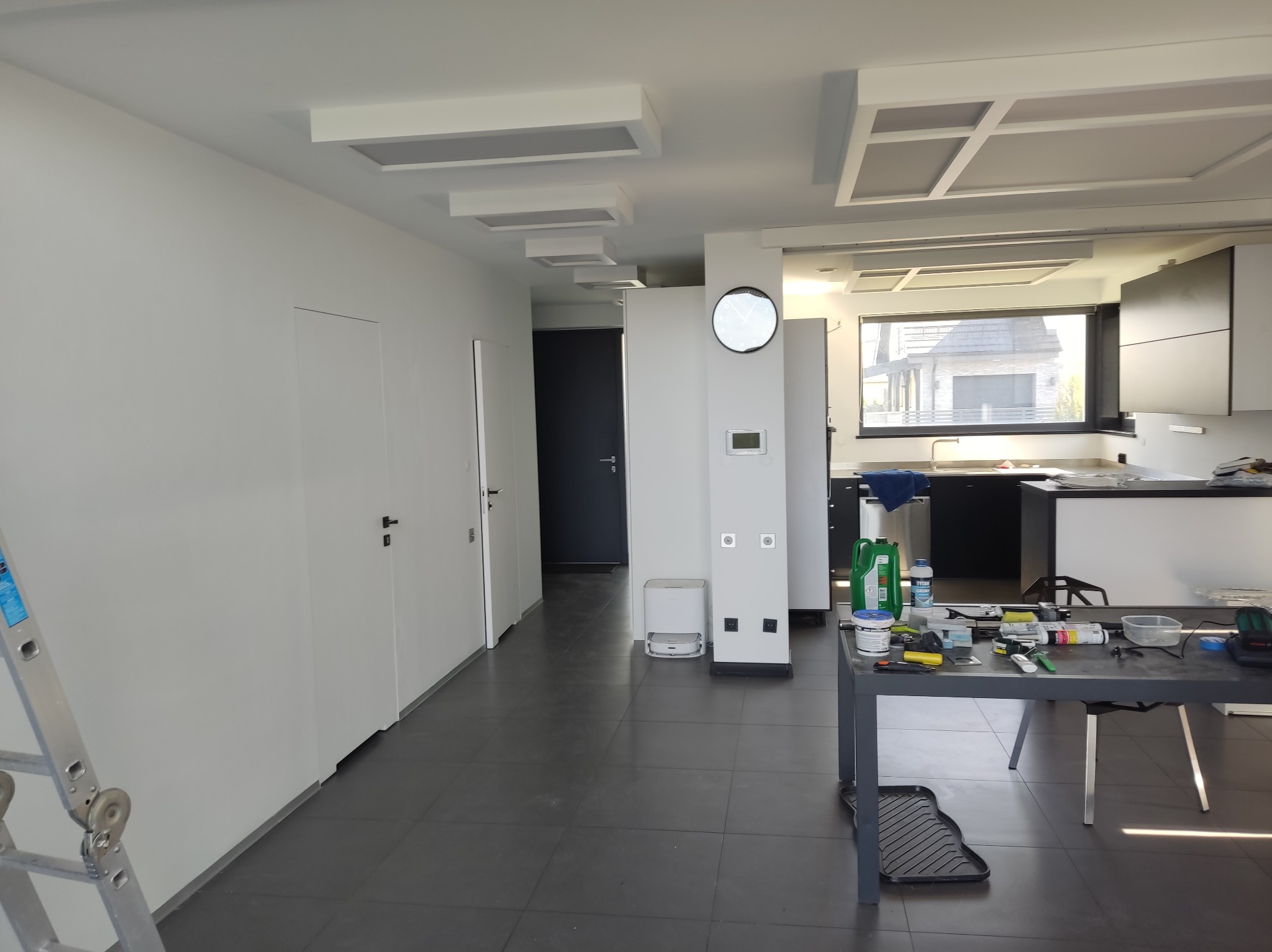 Wnętrze po malowaniu: białe ściany, sufit z panelami, ciemna podłoga. Widoczny stół z narzędziami malarskimi i drabina. Minimalistyczny design, nowoczesne oświetlenie.