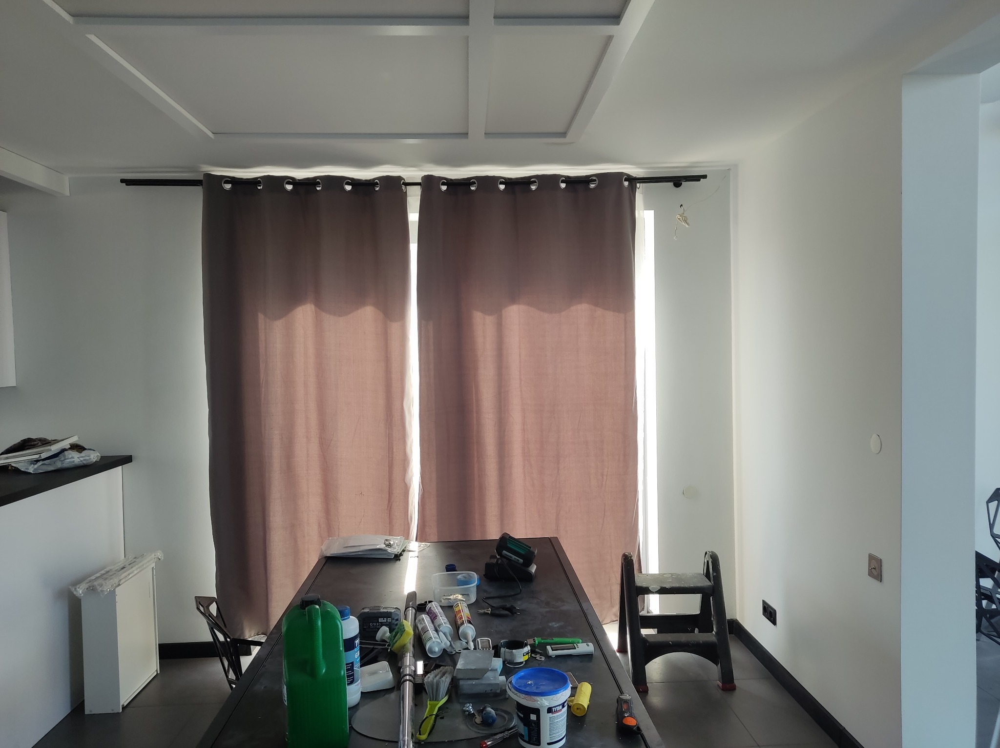 Przygotowania do malowania: stół z narzędziami, farbami i akcesoriami malarskimi w jasnym wnętrzu z zasłoniętymi oknami. Widoczna drabina i świeżo pomalowane ściany.