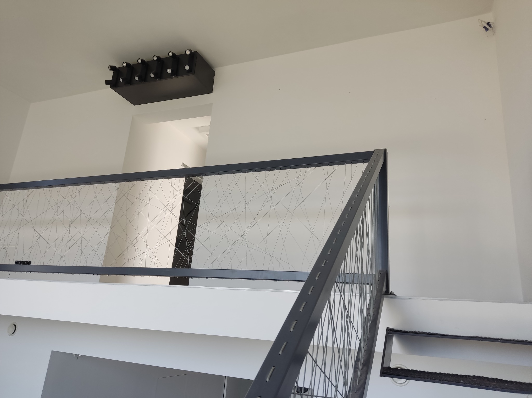 Minimalistyczne wnętrze z białymi ścianami, szarą balustradą z metalową siatką i nowoczesnym oświetleniem sufitowym w czarnej obudowie z wieloma punktami świetlnymi.