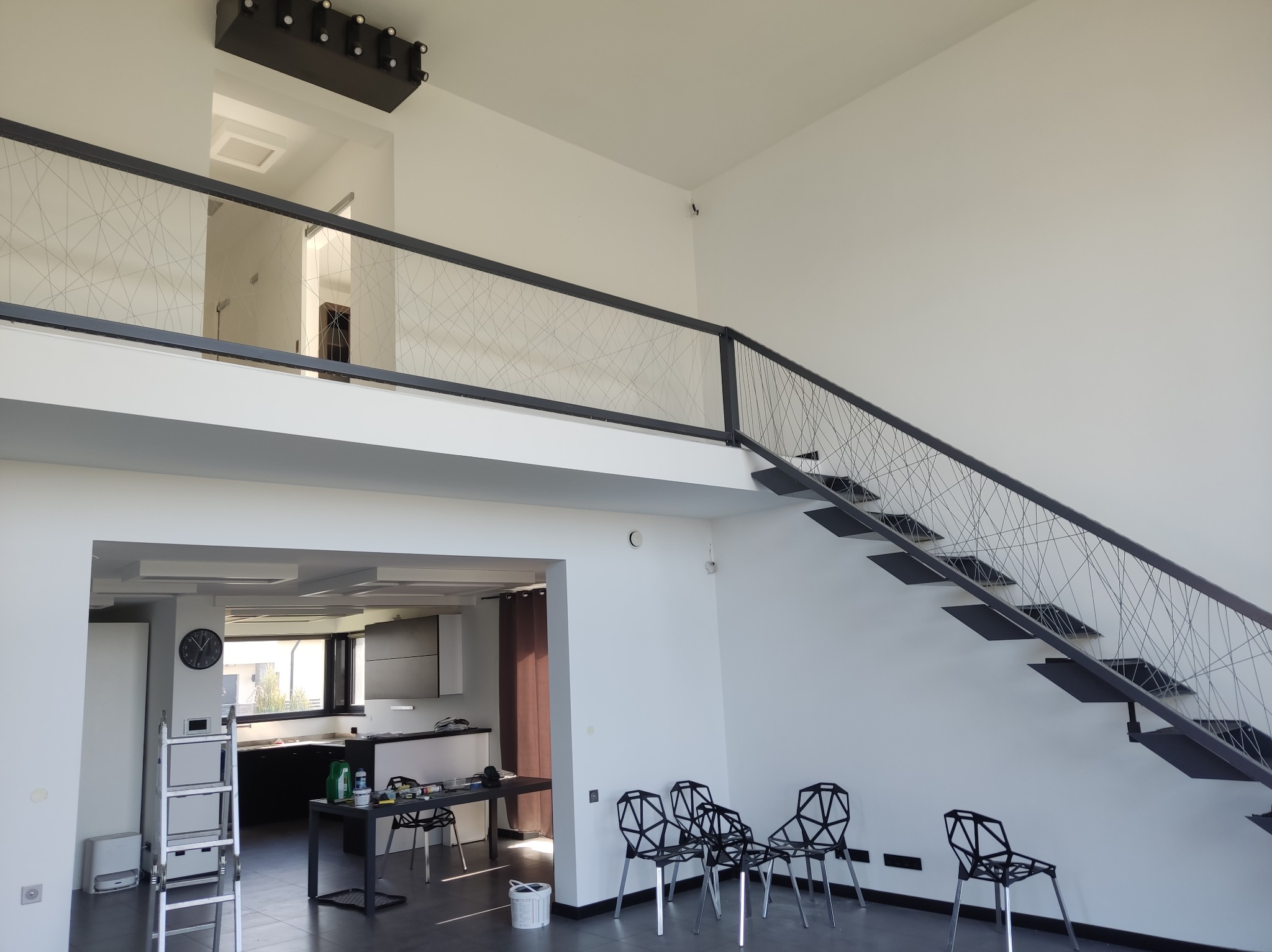 Wnętrze domu po malowaniu: białe ściany, schody z metalową balustradą, widoczna kuchnia. Na podłodze krzesła i akcesoria malarskie.