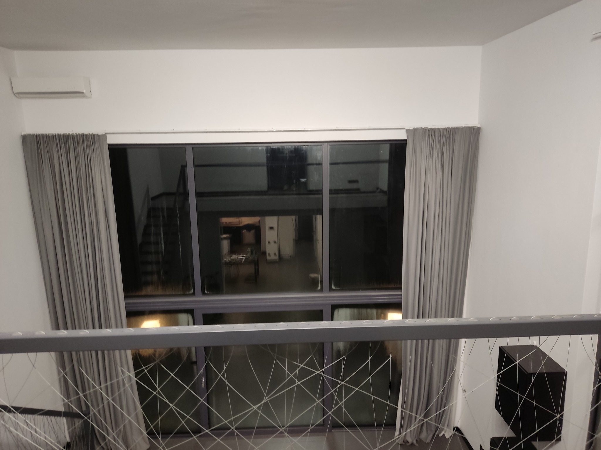 Wnętrze z dużym oknem i szarymi zasłonami, widok z góry przez metalową barierkę. Minimalistyczny design, jasne ściany, klimatyzator na ścianie. Widoczny fragment schodów.