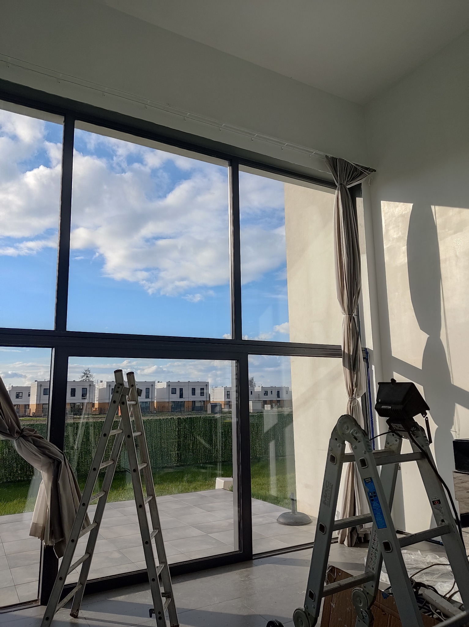 Montaż karnisza i zasłon w nowoczesnym wnętrzu z dużym oknem. Dwie drabiny ustawione przy oknie, zasłony częściowo zawieszone. Widok na osiedle domów.