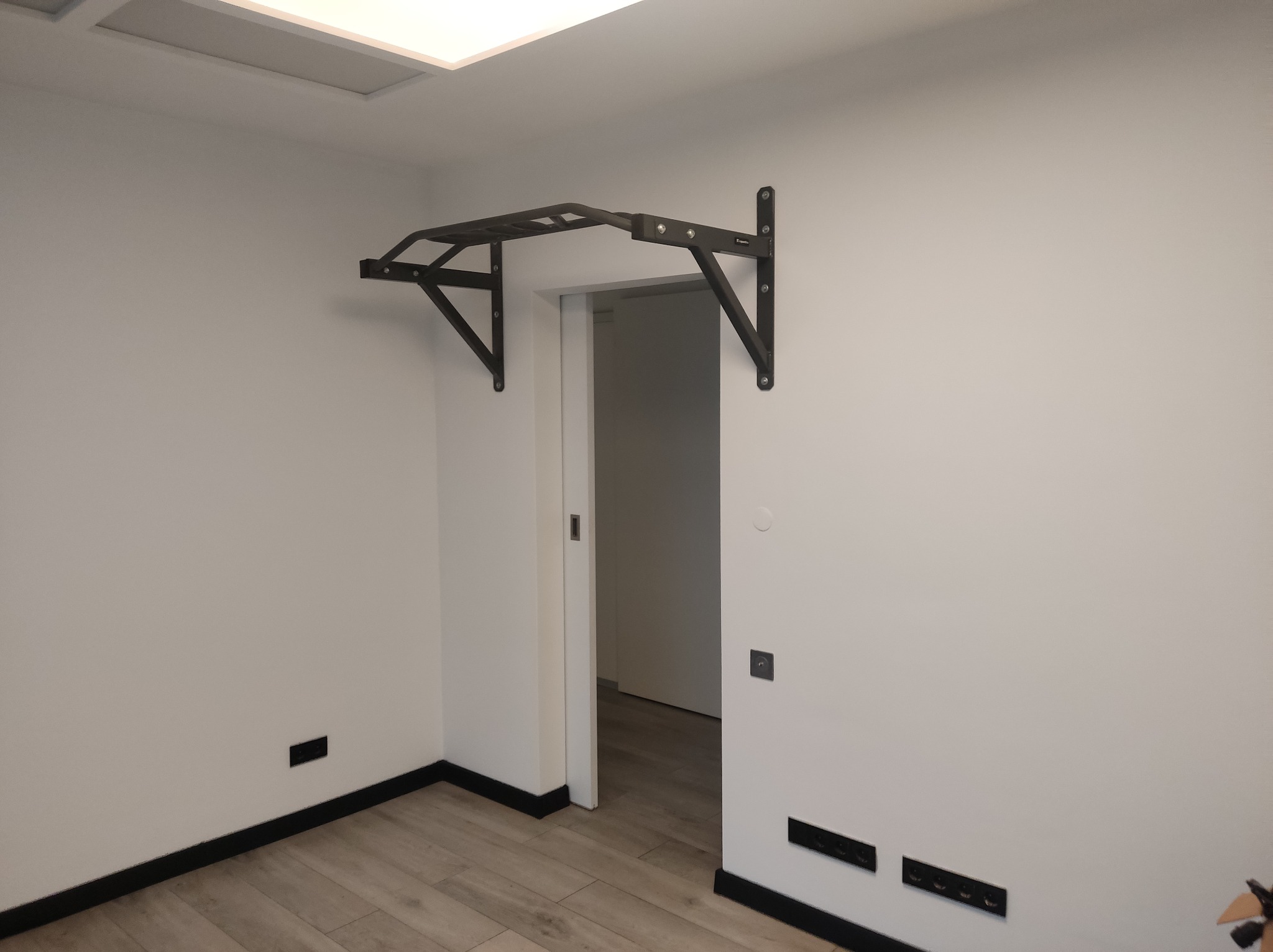 Czarny, metalowy drążek do podciągania zamontowany nad futryną drzwi w jasnym pokoju z drewnianą podłogą i czarnymi listwami przypodłogowymi.