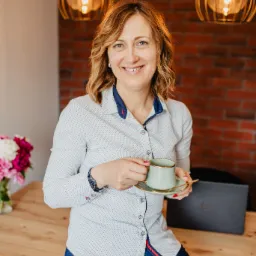 Uśmiechnięta kobieta w koszuli z wzorem, siedząca na drewnianym biurku z filiżanką kawy i złotą łyżeczką, na tle ceglanej ściany i lamp. W tle laptop i wazon z kwiatami.